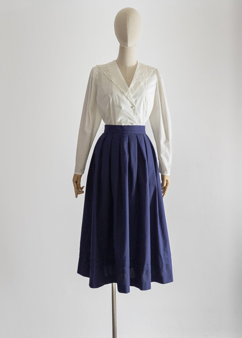 May include: A vintage outfit displayed on a mannequin. It comprises a white blouse with a lace collar and long sleeves, and a navy blue pleated midi skirt. The outfit is set against a plain white backdrop.