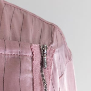 May include: Close-up of a pink garment with vertical black pinstripes. A silver zipper runs down the front, partially open. The fabric appears to be a satin or silk blend, with a slight sheen. The garment's construction is visible.