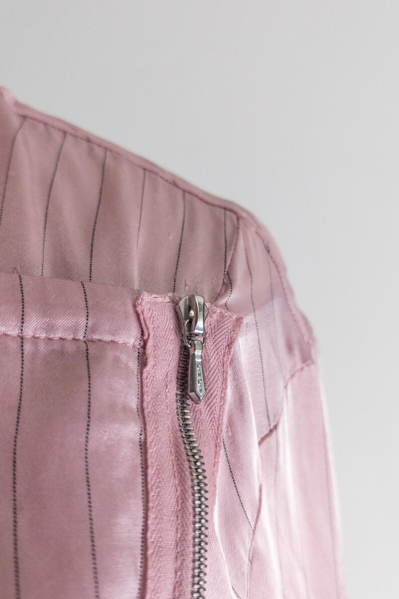 May include: Close-up of a pink garment with vertical black pinstripes. A silver zipper runs down the front, partially open. The fabric appears to be a satin or silk blend, with a slight sheen. The garment's construction is visible.