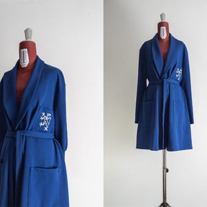 May include: A long, royal blue robe with a shawl collar, long sleeves, and a matching belt. The robe features two front pockets and a white embroidered floral design on the left side. The robe is displayed on a mannequin.