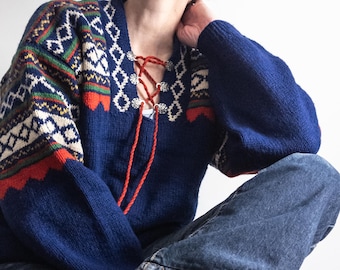 Könnte beinhalten: Blauer Pullover mit Muster auf Ärmeln und Brust. Der Pullover hat einen V-Ausschnitt mit roter Schnürung und silberfarbenen Verzierungen. Die Ärmel zeigen ein geometrisches Muster in Weiß, Grün und Rot.