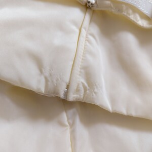 May include: Close-up of a white zipper on a garment. The zipper is partially open, revealing the white fabric underneath.