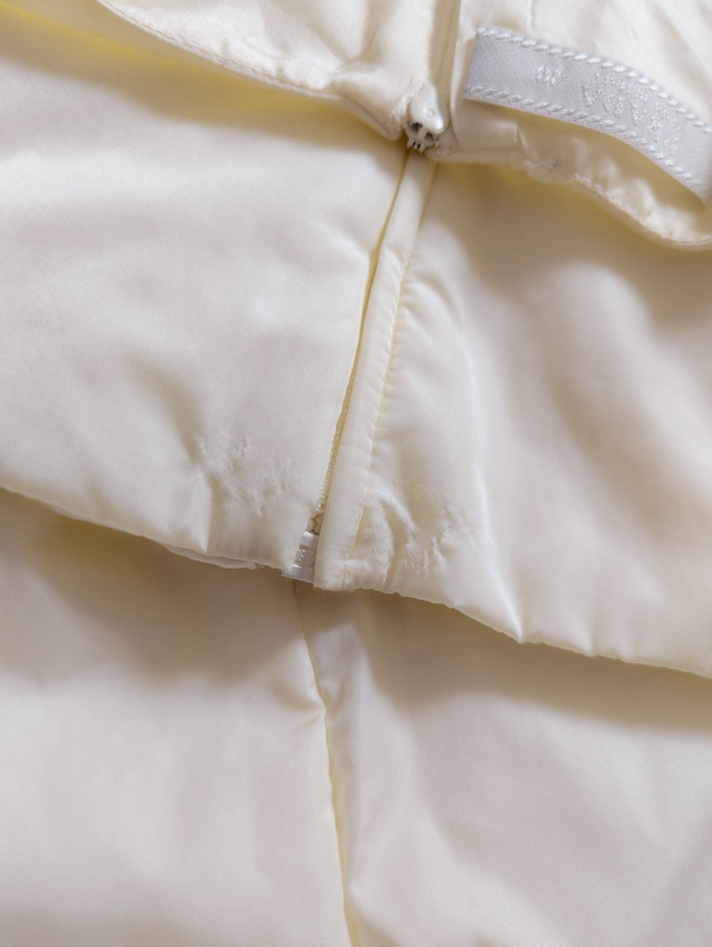 May include: Close-up of a white zipper on a garment. The zipper is partially open, revealing the white fabric underneath.