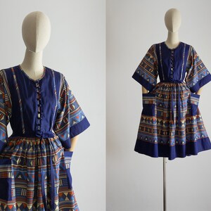 Könnte beinhalten: Ein blaues und orangefarbenes gemustertes Kleid mit Knopfleiste vorne, kurzen Ärmeln und Taschen. Das Kleid hat ein tailliertes Oberteil und einen weiten Rock.