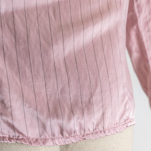 May include: Close-up of a pink, long-sleeved blouse with vertical black pinstripes. The fabric has a slight sheen, and the bottom hem is gathered. The sleeve is visible on the right side of the image.