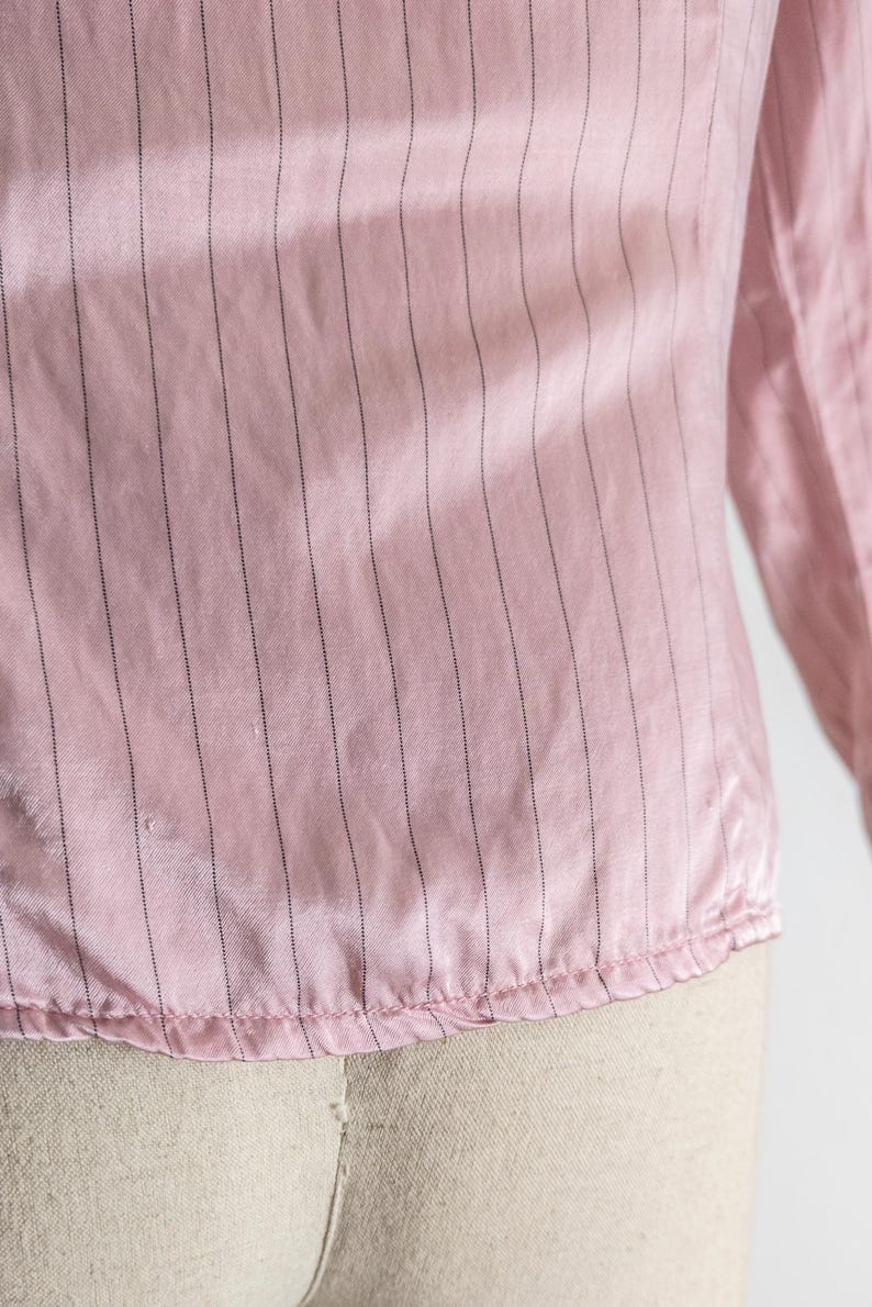 May include: Close-up of a pink, long-sleeved blouse with vertical black pinstripes. The fabric has a slight sheen, and the bottom hem is gathered. The sleeve is visible on the right side of the image.
