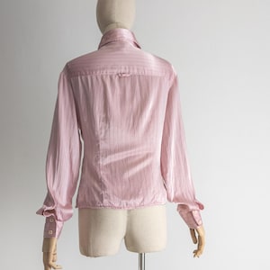 May include: A light pink, long-sleeved blouse with vertical stripes. The blouse features a collared neckline, buttoned cuffs, and a tailored fit. The back view of the blouse is displayed on a mannequin.