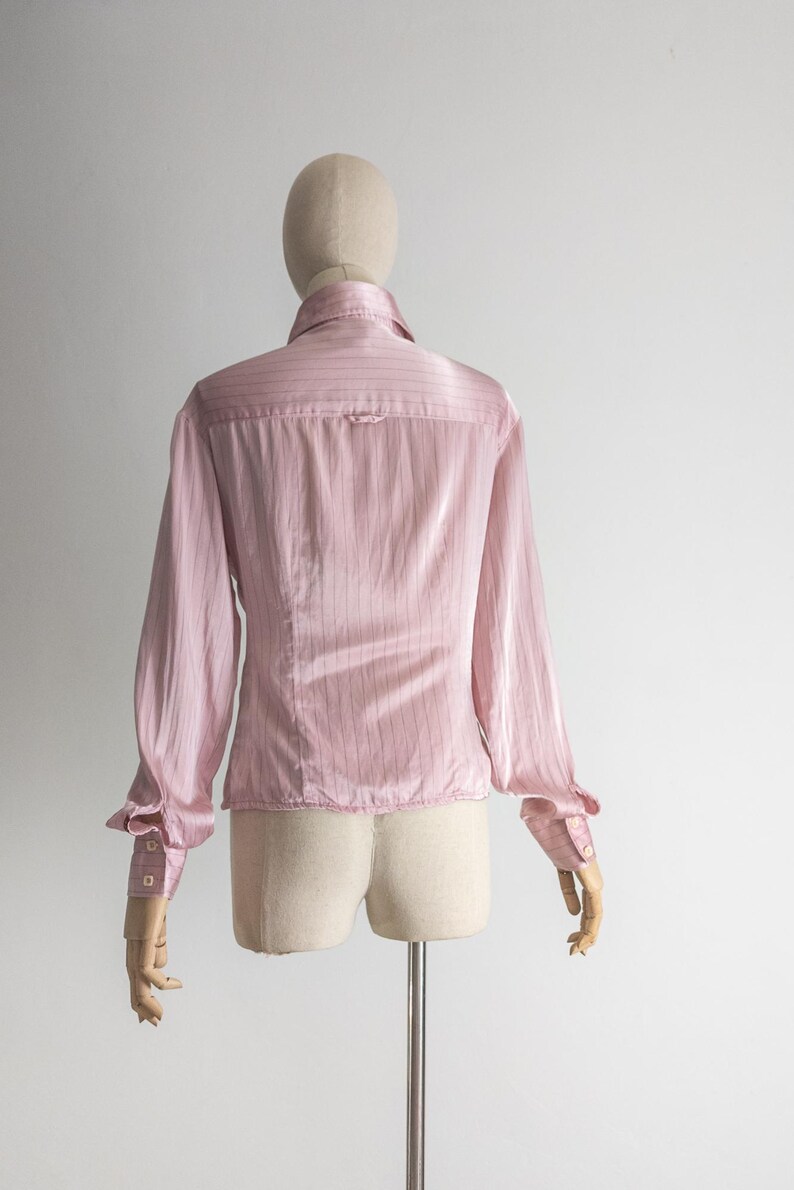May include: A light pink, long-sleeved blouse with vertical stripes. The blouse features a collared neckline, buttoned cuffs, and a tailored fit. The back view of the blouse is displayed on a mannequin.