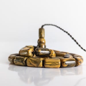Puede incluir: Primer plano de un juego de cuentas de oración. Las cuentas son una mezcla de amarillo, marrón y negro, con un efecto veteado. Las cuentas están ensartadas con un cordón negro, y una pieza decorativa a juego está en la parte superior.