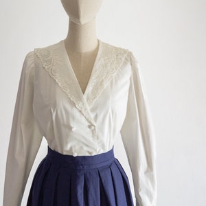 May include: A white blouse featuring a lace-trimmed collar and long sleeves, combined with a navy blue pleated skirt. The blouse has a double-breasted style with buttons. The outfit is displayed on a mannequin.