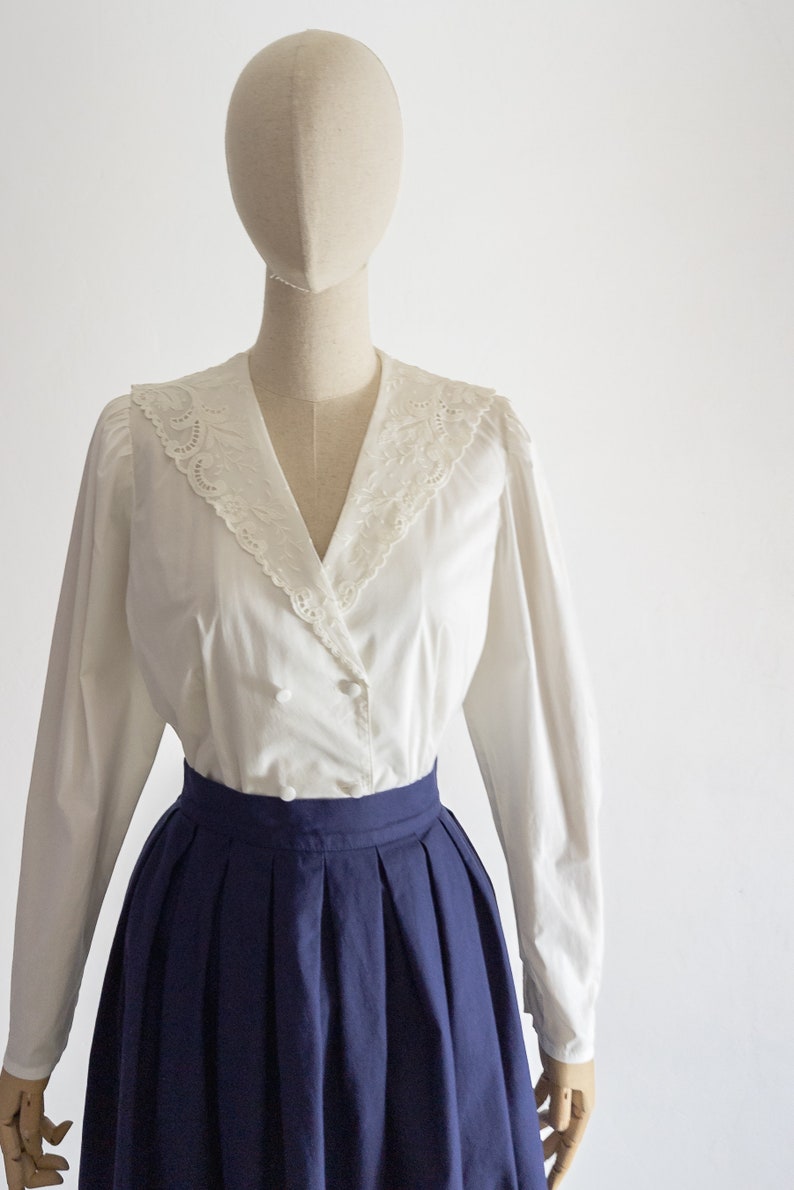 May include: A white blouse featuring a lace-trimmed collar and long sleeves, combined with a navy blue pleated skirt. The blouse has a double-breasted style with buttons. The outfit is displayed on a mannequin.