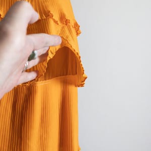 May include: A close-up of a bright orange dress with pleated fabric and ruffled tiers. The dress has a fitted bodice and a flowing skirt. The fabric appears lightweight and has a slight sheen. The dress is on a mannequin.