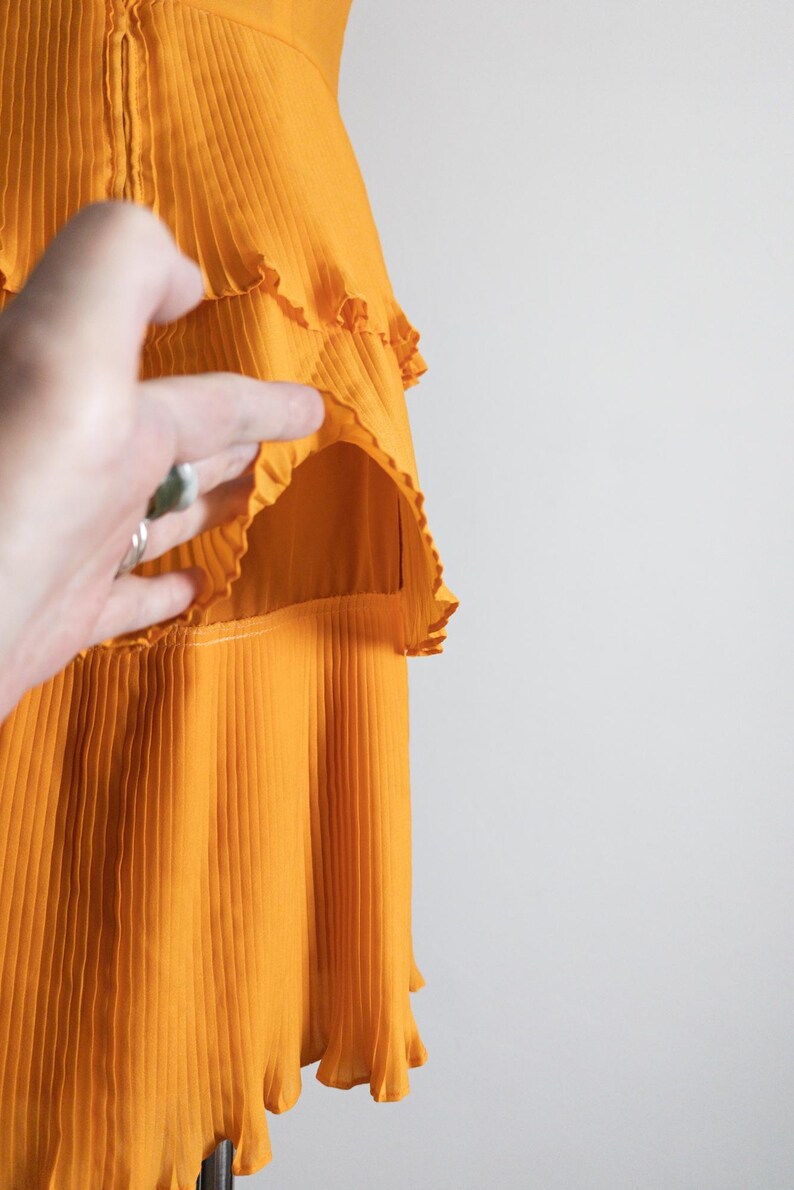 May include: A close-up of a bright orange dress with pleated fabric and ruffled tiers. The dress has a fitted bodice and a flowing skirt. The fabric appears lightweight and has a slight sheen. The dress is on a mannequin.