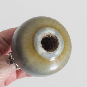 Puede incluir: Un pomo de puerta redondo y liso de cerámica en tonos verde oliva y gris, con una abertura circular central que revela un interior de metal roscado. La superficie del pomo presenta un esmalte moteado con finos patrones de grietas.