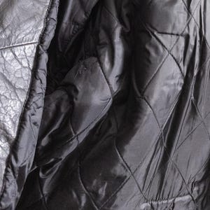 May include: Close-up of a black leather jacket interior. The lining is quilted with a diamond pattern. The jacket's exterior is visible on the left side, showing the texture of the leather. A label with text is visible on the upper right.