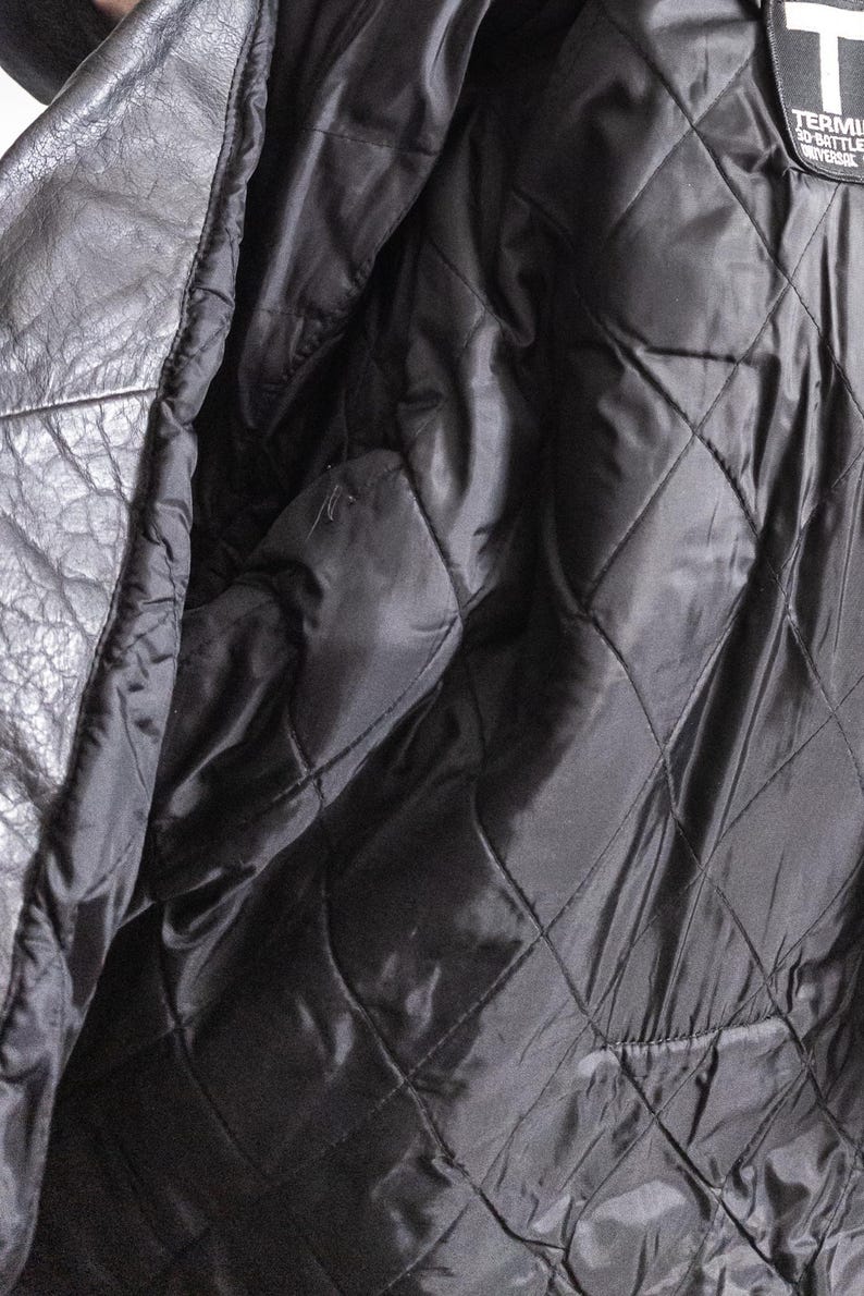 May include: Close-up of a black leather jacket interior. The lining is quilted with a diamond pattern. The jacket's exterior is visible on the left side, showing the texture of the leather. A label with text is visible on the upper right.