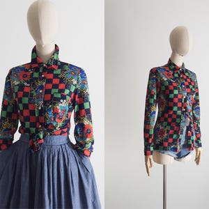 May include: A long-sleeved button-down shirt with a colorful checkerboard pattern of red, green, and black squares, interspersed with floral designs. The shirt is displayed on a mannequin, paired with a blue skirt and denim shorts.