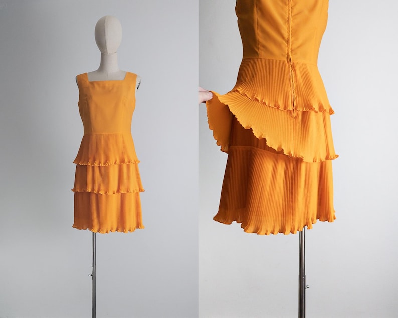 May include: A sleeveless, orange dress with a square neckline and a tiered, ruffled skirt. The dress is displayed on a mannequin. The fabric appears to be lightweight and has a slight sheen. The dress is a vibrant shade of orange.
