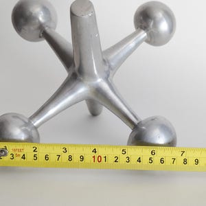 K&ouml;nnte beinhalten: Eine silberfarbene Metallskulptur in Form eines Spiel-Jacks mit sechs strahlenf&ouml;rmigen Armen, die in Kugeln enden. Ein gelbes Ma&szlig;band liegt davor und zeigt, dass das Objekt eine Breite von etwa 17,8 cm an den Au&szlig;enkanten der Kugeln hat.