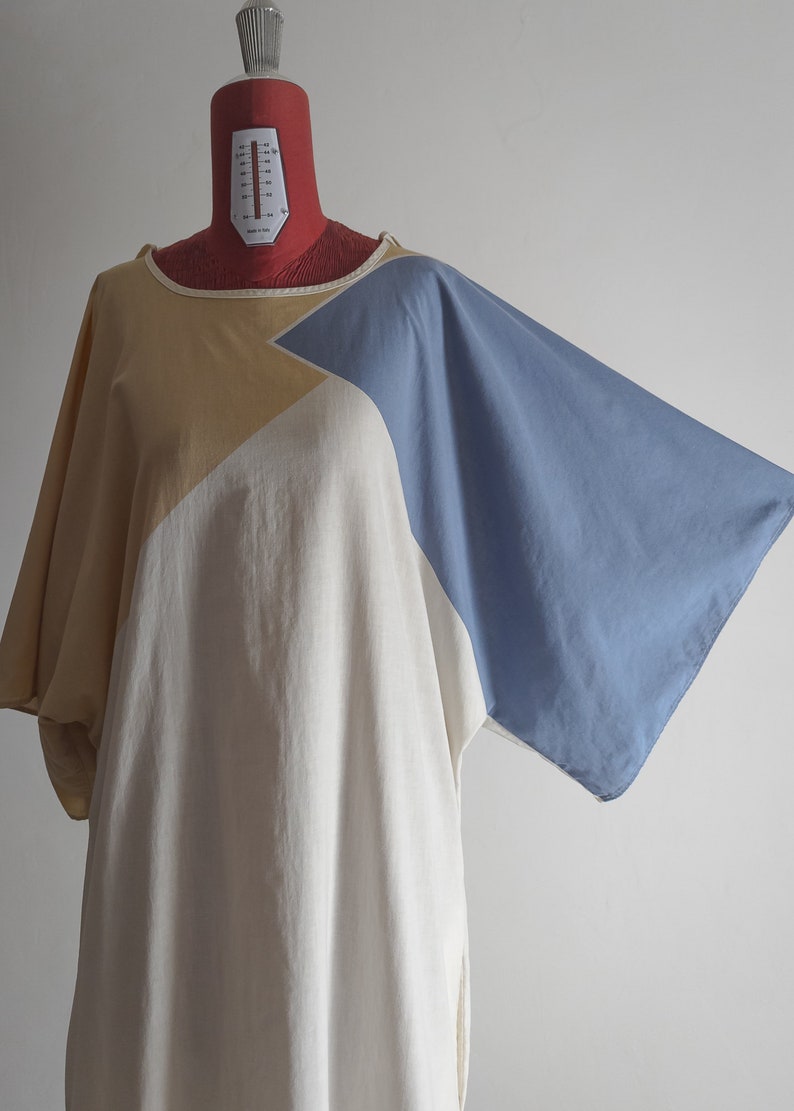 Op de afbeelding: Een loszittende, lange tuniekjurk met een geometrisch patroon in beige, wit en blauw. De jurk heeft een wijde, vloeiende silhouet en een ronde hals.