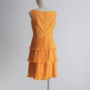 May include: A sleeveless, orange dress with a square neckline and a tiered, pleated skirt. The dress is displayed on a mannequin against a plain white background. The back of the dress features a zipper closure.