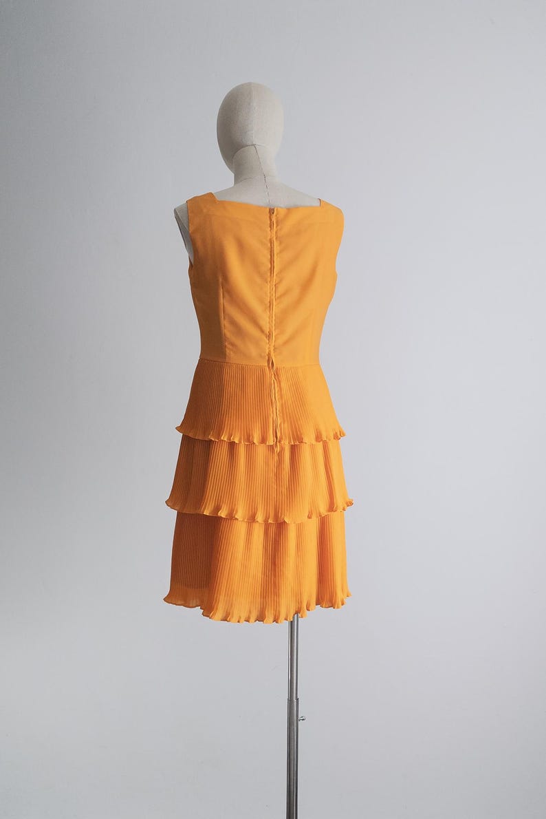 May include: A sleeveless, orange dress with a square neckline and a tiered, pleated skirt. The dress is displayed on a mannequin against a plain white background. The back of the dress features a zipper closure.