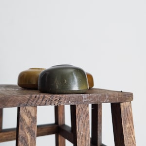 May include: Three small, rounded, wooden objects in shades of olive green and gold sit on a rustic wooden stool. The stool has a dark, aged appearance with visible wood grain. The background is a plain white wall.