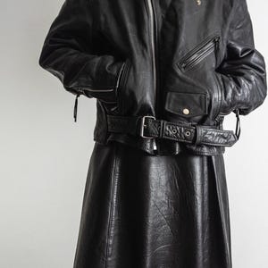 May include: Black leather jacket with a belted waist and a matching black leather skirt. The jacket features a zipper closure, multiple pockets, and shoulder epaulets. The skirt is a flared style.