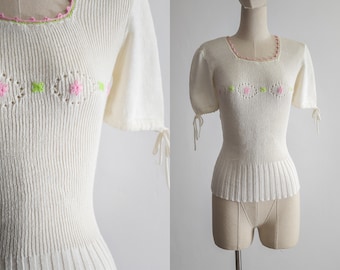 May include: White knitted sweater with short sleeves and a pink and green floral design. The sweater has a ribbed texture and a slightly fitted silhouette.