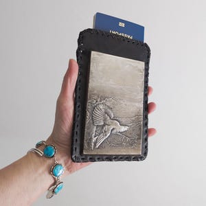 May include: Black leather passport holder with a decorative silver panel featuring a bird in flight. The holder has a stitched border and a passport is visible inside. A turquoise bracelet is visible in the background.