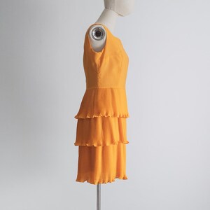 May include: A sleeveless, orange dress with a tiered, pleated skirt. The dress is displayed on a mannequin against a white background. The dress has a fitted bodice and a three-tiered skirt.