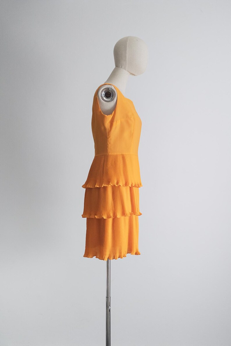 May include: A sleeveless, orange dress with a tiered, pleated skirt. The dress is displayed on a mannequin against a white background. The dress has a fitted bodice and a three-tiered skirt.