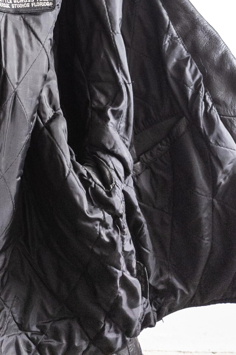 May include: Close-up of a black leather jacket interior. The quilted lining is visible, along with the smooth, shiny fabric of the jacket's interior. The jacket appears to be well-worn.