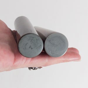 Puede incluir: Dos objetos cilíndricos con un acabado mate de color gris oscuro liso. Cada cilindro presenta una cara final circular plana con una textura veteada de color gris ligeramente más claro. Los objetos son alargados y de aspecto sólido.