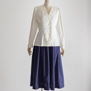 May include: A vintage outfit on a mannequin. The ensemble includes a white, long-sleeved blouse with a V-neck, lace detailing, and button closure. It is paired with a navy blue, knee-length skirt. The outfit is displayed on a mannequin.