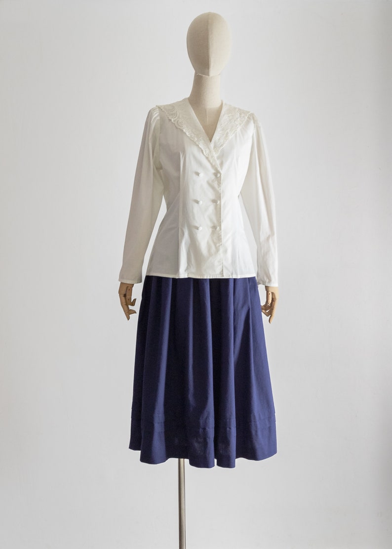 May include: A vintage outfit on a mannequin. The ensemble includes a white, long-sleeved blouse with a V-neck, lace detailing, and button closure. It is paired with a navy blue, knee-length skirt. The outfit is displayed on a mannequin.