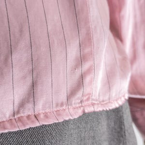 May include: Close-up of a pink blouse with vertical black pinstripes. The fabric has a slight sheen and a ruffled hem. The bottom portion of the image shows a gray fabric.