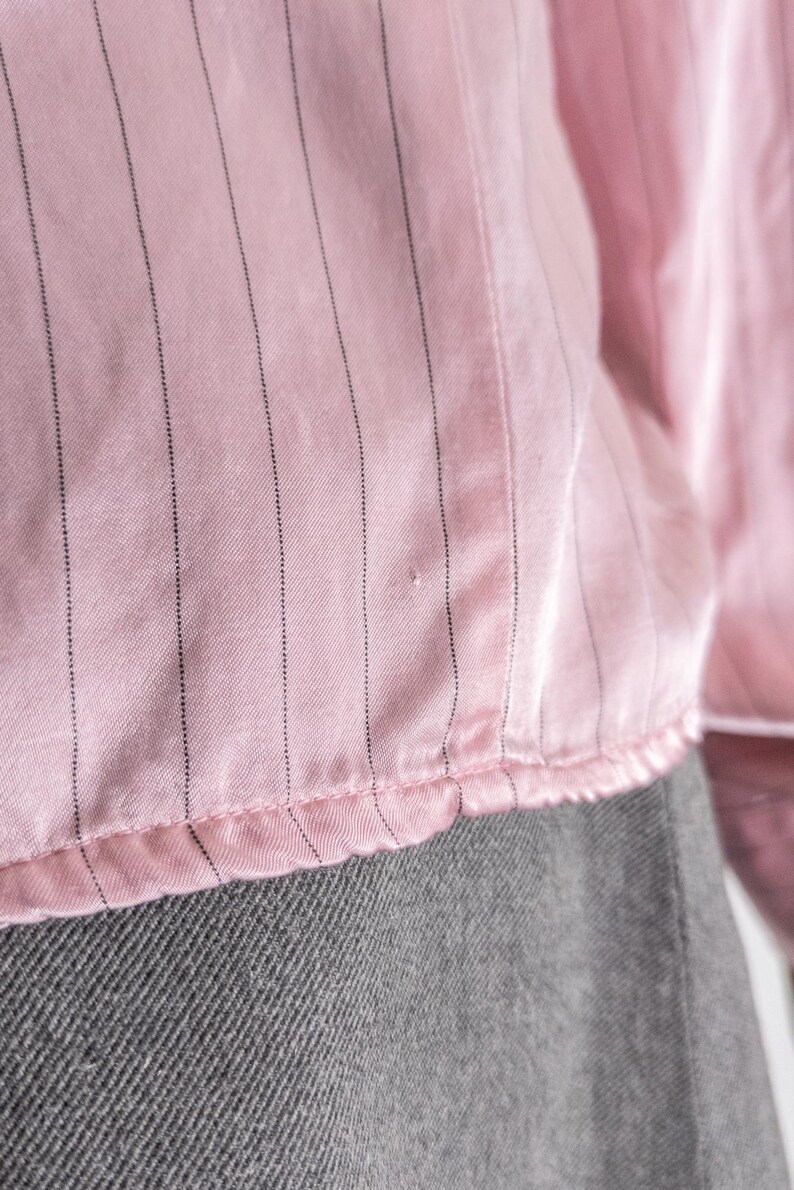 May include: Close-up of a pink blouse with vertical black pinstripes. The fabric has a slight sheen and a ruffled hem. The bottom portion of the image shows a gray fabric.