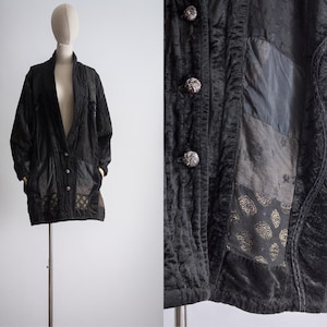 May include: A black quilted jacket with a patchwork design featuring different shades of gray and a gold pattern. The jacket has a button closure and a collar.