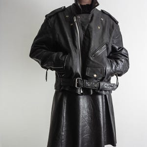 May include: Black leather jacket with a belted waist, multiple pockets, and a zipper closure. The jacket has shoulder epaulets and a classic biker style. The matching black leather skirt is pleated.