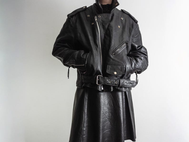 May include: Black leather jacket with a belted waist, multiple pockets, and a zipper closure. The jacket has shoulder epaulets and a classic biker style. The matching black leather skirt is pleated.