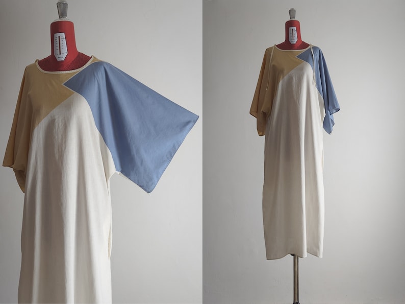 Op de afbeelding: Een losse, wit, geel en blauwe kleurblokjurk met korte mouwen. De jurk heeft een geometrisch ontwerp met een grote driehoekvorm aan de voorkant.