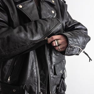 May include: Black leather motorcycle jacket with silver zippers, snap closures, and a notched collar. The jacket has a classic biker style with a belted waist and multiple pockets. The leather appears worn, giving it a vintage look.
