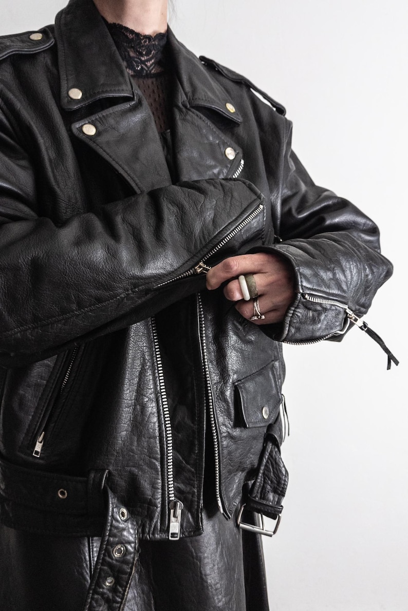 May include: Black leather motorcycle jacket with silver zippers, snap closures, and a notched collar. The jacket has a classic biker style with a belted waist and multiple pockets. The leather appears worn, giving it a vintage look.