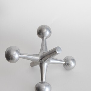 K&ouml;nnte beinhalten: Eine silberfarbene Metallskulptur in Form eines Spielzeug-Jacks, bestehend aus einem zentralen Knotenpunkt mit sechs strahlenf&ouml;rmigen Armen, die jeweils in einer glatten, kugelf&ouml;rmigen Kugel enden. Das Objekt hat eine polierte Oberfl&auml;che.
