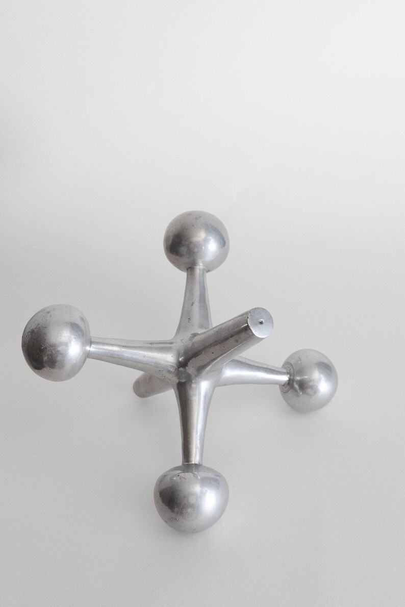 K&ouml;nnte beinhalten: Eine silberfarbene Metallskulptur in Form eines Spielzeug-Jacks, bestehend aus einem zentralen Knotenpunkt mit sechs strahlenf&ouml;rmigen Armen, die jeweils in einer glatten, kugelf&ouml;rmigen Kugel enden. Das Objekt hat eine polierte Oberfl&auml;che.