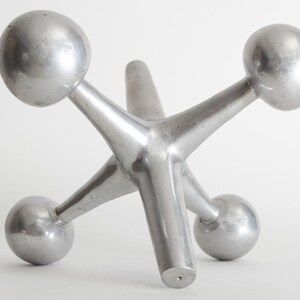 K&ouml;nnte beinhalten: Eine silberfarbene Metallskulptur in Form eines Spiel-Jacks mit sechs strahlenf&ouml;rmigen Armen, die jeweils in einer glatten Kugel enden. Das Objekt hat eine polierte, reflektierende Oberfl&auml;che und eine solide, dreidimensionale Struktur.