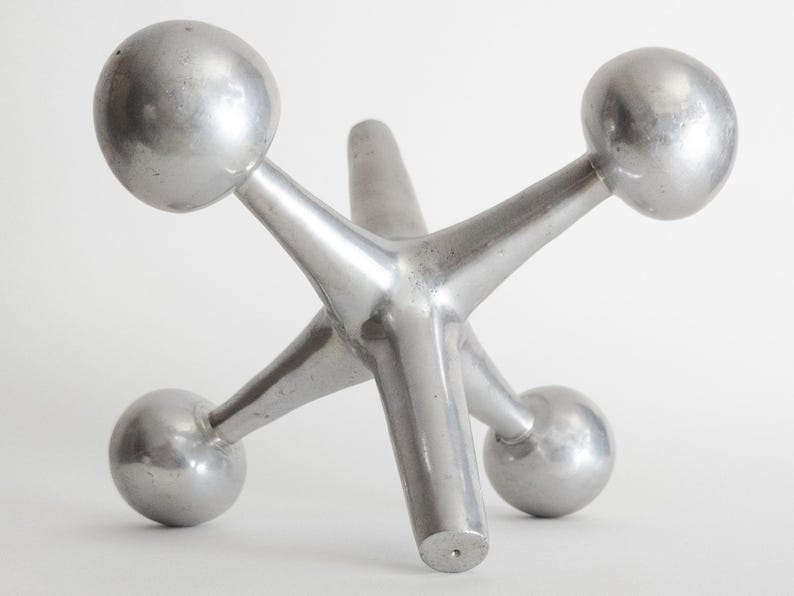 K&ouml;nnte beinhalten: Eine silberfarbene Metallskulptur in Form eines Spiel-Jacks mit sechs strahlenf&ouml;rmigen Armen, die jeweils in einer glatten Kugel enden. Das Objekt hat eine polierte, reflektierende Oberfl&auml;che und eine solide, dreidimensionale Struktur.