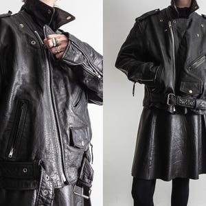 May include: Black leather jacket with a high collar, shoulder epaulets, and multiple pockets. The jacket features a silver zipper and a belted waist. The model is wearing a black leather skirt and black leggings.