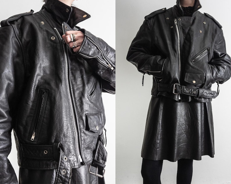 May include: Black leather jacket with a high collar, shoulder epaulets, and multiple pockets. The jacket features a silver zipper and a belted waist. The model is wearing a black leather skirt and black leggings.